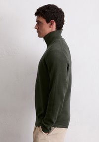 Ciemnozielony, dzianinowy sweter z wysokim kołnierzem i półzamkiem. Dopasowany krój, z prążkowanymi mankietami i dołem. Gładka tekstura, minimalistyczny design.
