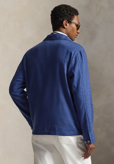 Άντρας που φορά μπλε μακρυμάνικο μπουφάν και λευκό παντελόνι, με την πλάτη στραμμένη, με κοντά σγουρά μαλλιά και σκοτεινά γυαλιά ηλίου σε απλό φόντο.