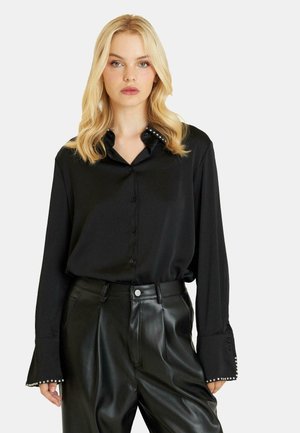 Camisa de satén negro con botones, cuello con acentos de perlas y mangas largas. Combinada con pantalones de piel negra de cintura alta.