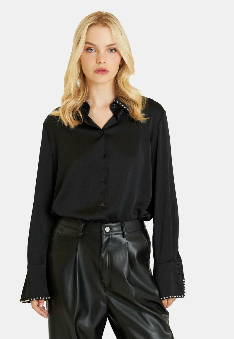 Camisa de satén negro con botones, cuello con acentos de perlas y mangas largas. Combinada con pantalones de piel negra de cintura alta.