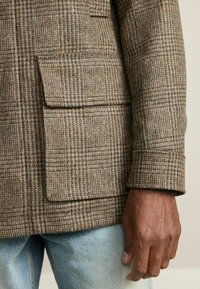 Pruun ruuduline mantel, mis on valmistatud tekstuuriga kangast, suure küljetaskuga ja üleskeeratud varrukatega. Kandes helesiniste teksade peal.