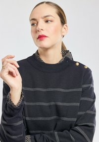 Maglione blu navy con strisce bianche orizzontali, colletto rotondo a costine e dettagli con bottoni dorati sulle spalle, indossato sopra una camicia a quadri.