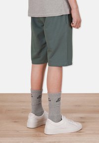 Gröna shorts i ett mjukt material, åtsittande vid midjan med sidfickor, i kombination med grå strumpor och vita sneakers på ett trägolv.