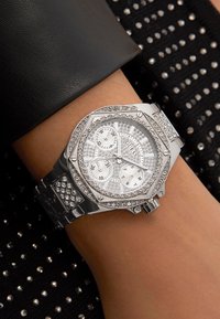 Montre en argent avec un cadran octogonal, plusieurs compteurs et des accents sertis. Bracelet en métal argenté avec des détails scintillants, portée sur un poignet contre un tissu noir.