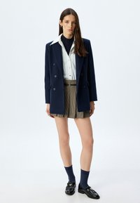 Blazer doppiopetto blu navy sopra una camicia bianca, abbinato a pantaloni corti a scacchi, calzini blu navy e mocassini neri con dettagli dorati.