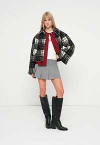 Μαύρο και άσπρο καρό cropped σακάκι, κόκκινο πουλόβερ με κουμπιά, λευκό T-shirt, γκρι πλισέ φούστα και μαύρες μπότες μέχρι το γόνατο.