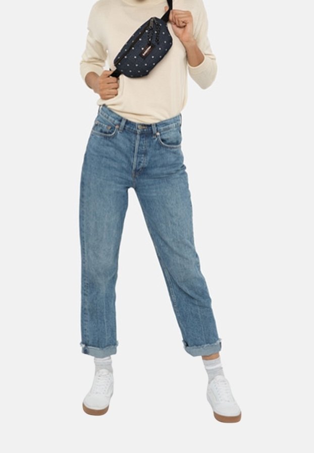 Personne portant un jean bleu roulé, des baskets blanches, des chaussettes claires et une chemise à manches longues claire, tenant un sac bandoulière bleu marine avec un motif.