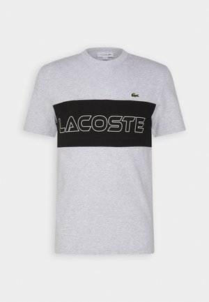T-shirt gris clair à manches courtes avec une bande horizontale noire sur la poitrine comportant le texte "LACOSTE" en contour blanc et un petit logo crocodile vert.