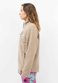 Beige Fleecejacke mit hohem Kragen, Frontreißverschluss und Seitentaschen. Strukturiertes Material und ein kleines Logopatch auf der Brust.