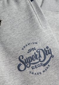 Tejido de algodón gris con un gran logo bordado en azul marino que presenta el texto "SuperDry" y detalles adicionales de la marca.
