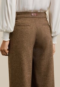 Pantalons à carreaux houndstooth marron à jambes larges avec une texture lisse. Comprend des poches arrière et une étiquette en cuir de marque. Associés à un doux chemisier blanc.