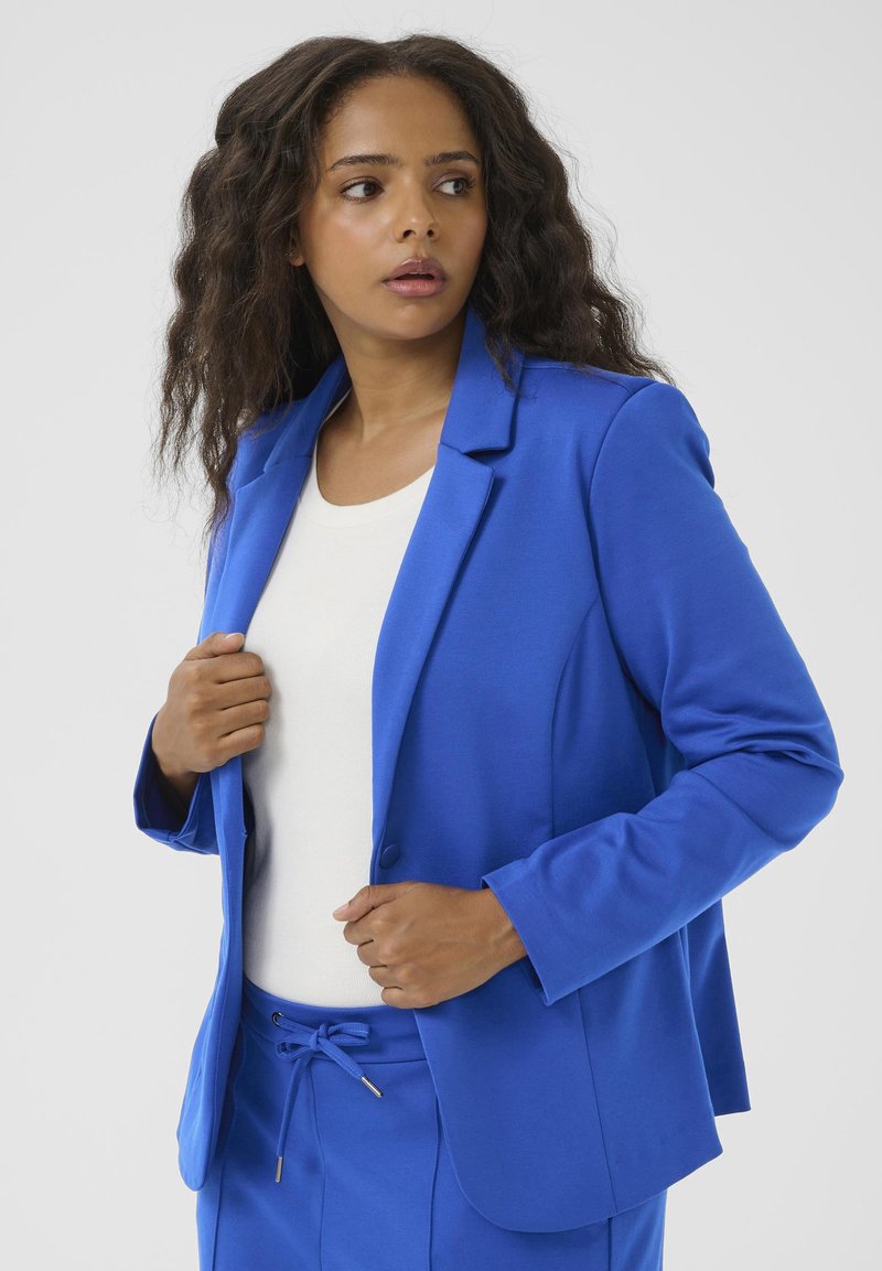 Blauwe blazer van een gladde stof met een getailleerde pasvorm, sluiting met één knop en notch revers, gecombineerd met een witte top.