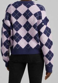 Stickad tröja i lila och marinblå rutig mönster, med kontrasterande vita linjer. Har knappdetaljer på ärmarna och en ribbad nederkant.