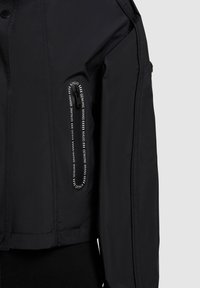 Manche d'une veste noire avec une poche zippée verticale bordée de texte blanc indiquant "KHUJO | GENUINE BRAND" répété autour de la fermeture éclair.