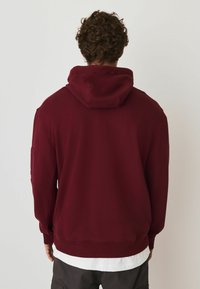 Bordeauxrode hoodie met een grote capuchon, geribbelde manchetten en tailleband. Gemaakt van zacht materiaal, met een gladde textuur en een minimalistisch ontwerp.