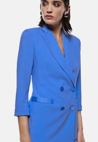 Blazer blu doppiopetto con rever, dotato di bottoni in tessuto, rifiniture in raso sulle tasche e maniche a tre quarti. Texture liscia.