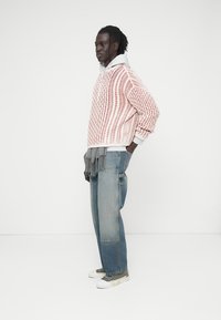 Homme portant un pull en maille rose et blanc par-dessus un sweat à capuche blanc, un jean large patchwork, une écharpe grise et des baskets grises, debout de profil.