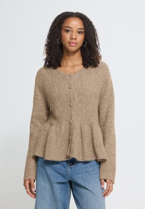 Frau trägt eine beigefarbene Strickpeplumjacke mit Knopfleiste vorne und blaue Jeans, steht vor einem hellen, schlichten Hintergrund und blickt nach vorne.