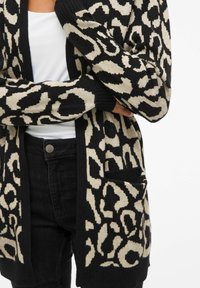 Kardigan z leopardjim vzorjem v bež in črni barvi, s sproščenim krojem, dolgimi rokavi in prednjim žepom, izdelan iz mehkega pletenega materiala.