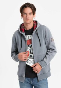 Felpa grigia con zip, con un rivestimento a quadri rossi dentro il cappuccio, presenta una tasca frontale, lacci e polsini a coste. Indossata sopra una maglietta nera con grafica.