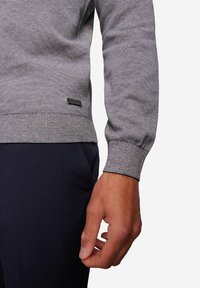 Gestrickter Pullover in grauen Streifen mit einem strukturierten Muster, ausgestattet mit einem gerippten Bündchen und einem schwarzen Logo-Patch an der Taille. Dunkle Hosen sind sichtbar.