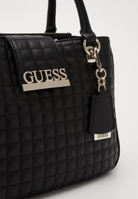 Černá prošívaná kabelka s zlatým logem "GUESS" na kovovém štítku, přívěskem a texturovaným povrchem. Vlastnosti zahrnují dvojité uchycení a zipové zavírání.