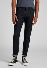 Jeans de mezclilla azul oscuro con un ajuste slim y una textura sutil. Llevados con una camisa azul marino y zapatillas negras de caña alta, con los puños arremangados.