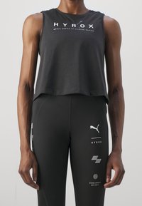Top atletico nero cropped con logo bianco "HYROX". Indossato con leggings neri che mostrano vari grafici legati al fitness e il logo Puma.