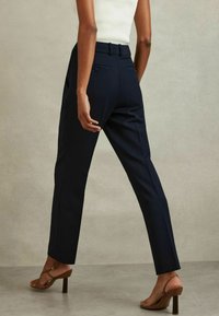 Žena nosící tmavě modré kalhoty na míru do kotníků, bílý tílko bez rukávů a hnědé sandály na podpatku, stojící před jednobarevným neutrálním pozadím.