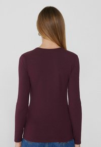 Burgunderfarbener Langarmoberteil mit rundem Ausschnitt, taillierter Form und glatter Textur, von hinten mit minimaler Detailgestaltung gezeigt.