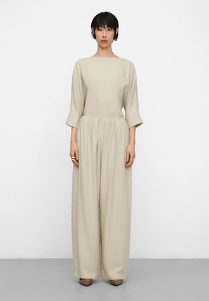 Donna che indossa una tuta beige lunga e ampia con maniche a tre quarti, in piedi davanti a uno sfondo bianco semplice.