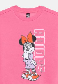 Ružový crewneck sveter s grafikou karikatúrnej postavy s červenou mašľou, fialovým oblečením a oranžovými teniskami s bielymi detailmi na podrážke.
