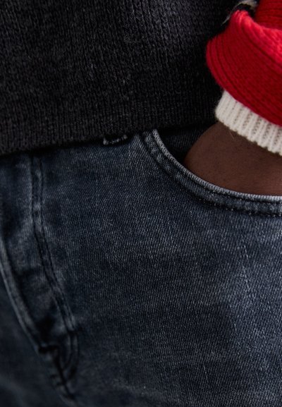 Jeans in denim grigio scuro con una texture sottile, caratterizzati da una tasca posteriore e una mano parzialmente visibile. Un maglione a maglia nera si trova sopra.