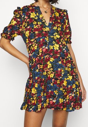 Žena v krátkých rukávech květinových šatech s žlutými, červenými, modrými a růžovými květy na černém pozadí a zlatým přívěskem na náhrdelníku.