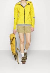 Keltainen vedenpitävä takki harmailla yksityiskohdilla, beiget shortsit ja keltainen reppu. Ruskeat ja vihreät kengät, valkoiset sukat. Yksinkertainen malli, rento istuvuus.