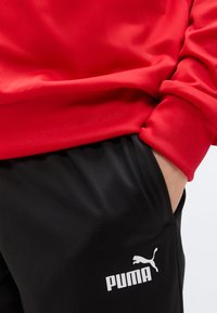 Κόκκινο φούτερ με απαλή υφή, συνδυασμένο με μαύρο φόρμας παντελόνι με λευκό λογότυπο Puma. Το παντελόνι έχει άνετη εφαρμογή.