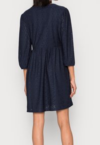 Robe bleu marine avec des manches bouffantes trois-quarts, présentant un motif texturé en œillets et une taille cintrée, se déversant en une jupe décontractée.