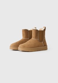 Pair of tan suede ankle boots with thick rubber soles and white fleece lining, facing slightly right on a plain white background.