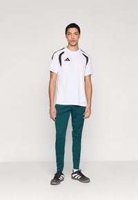 Vyrams skirtas sportinis aprangos komplektas: balti trumpomis rankovėmis marškinėliai su juodais elementais, žaliai prigludusios kelnės ir juodi batai su baltais dryžiais.