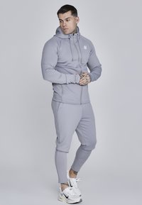 Sivi sportski dukserica sa zatvaračem, kengur džepom i logom; uparen sa odgovarajućim sivim suženim hlačama i bijelim tenisicama.