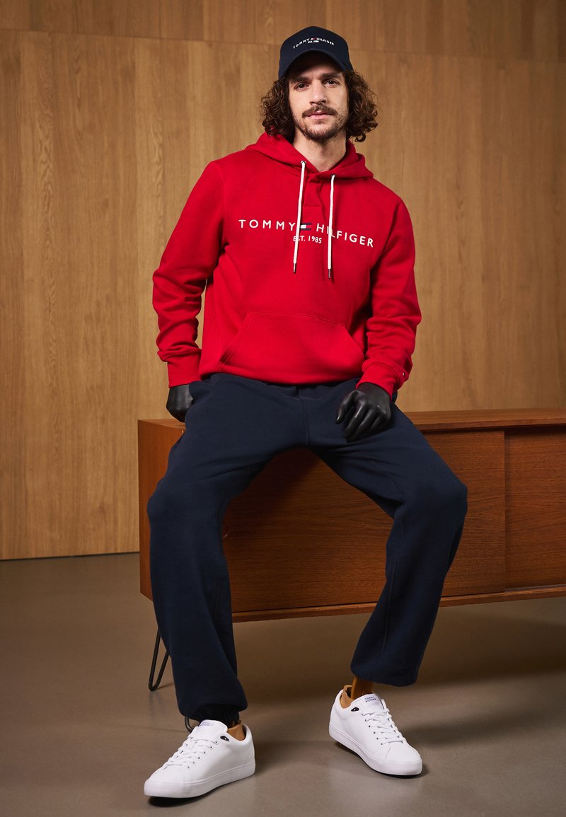 Rød hettegenser med hvitt logo, marineblå joggebukser og hvite joggesko. Hettegenseren har kengurulomme og justerbare snorer.