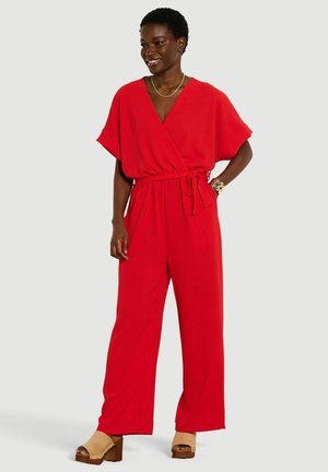 Vrouw draagt een rode wrapstijl jumpsuit met korte mouwen, een taillekoord, beige platformsandalen en gouden sieraden, glimlachend en poserend.