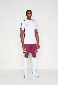 Férfi áll fehér és fekete Nike sportpólóban, bordó rövidnadrágban, fehér térdig érő zoknival és világoskék Nike stoplis cipőben, egyszínű háttér előtt.
