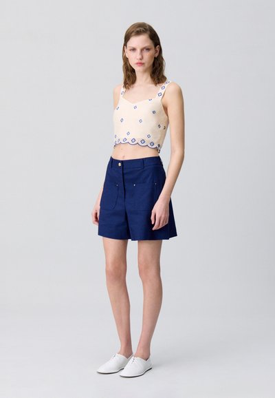 Haut court beige avec détail de broderie bleue, associé à un short marine avec deux poches avant et une fermeture par bouton. Chaussures blanches.