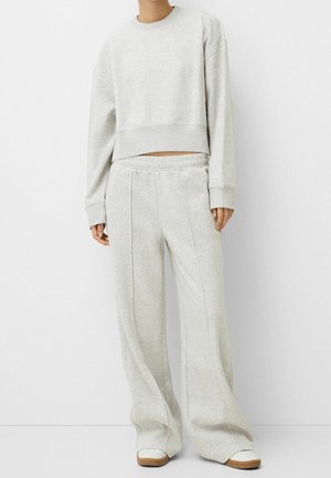 Osoba ubrana w jasnoszarą, oversize'ową bluzę oraz pasujące do niej szerokie spodnie dresowe wraz z białymi trampkami na jednolitym białym tle.