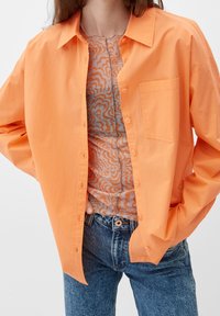 Orange skjorte med knapper og brystlomme, lagdelt over en mønstret top. Sammen med blå højtaljede jeans. Glat tekstur, afslappet pasform.
