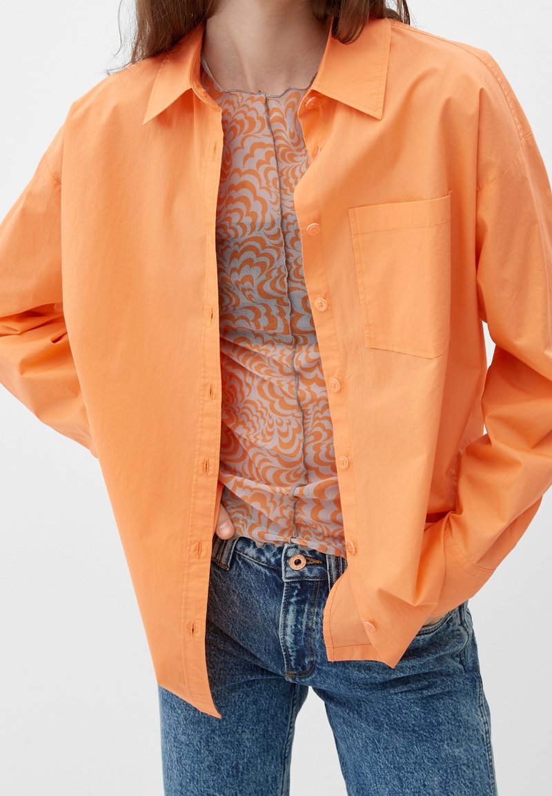 Orange skjorte med knapper og brystlomme, lagdelt over en mønstret top. Sammen med blå højtaljede jeans. Glat tekstur, afslappet pasform.