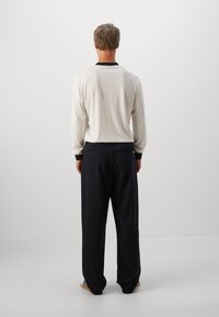 Homme de dos, portant une chemise à manches longues crème, un pantalon large noir et des baskets blanches avec des rayures noires.