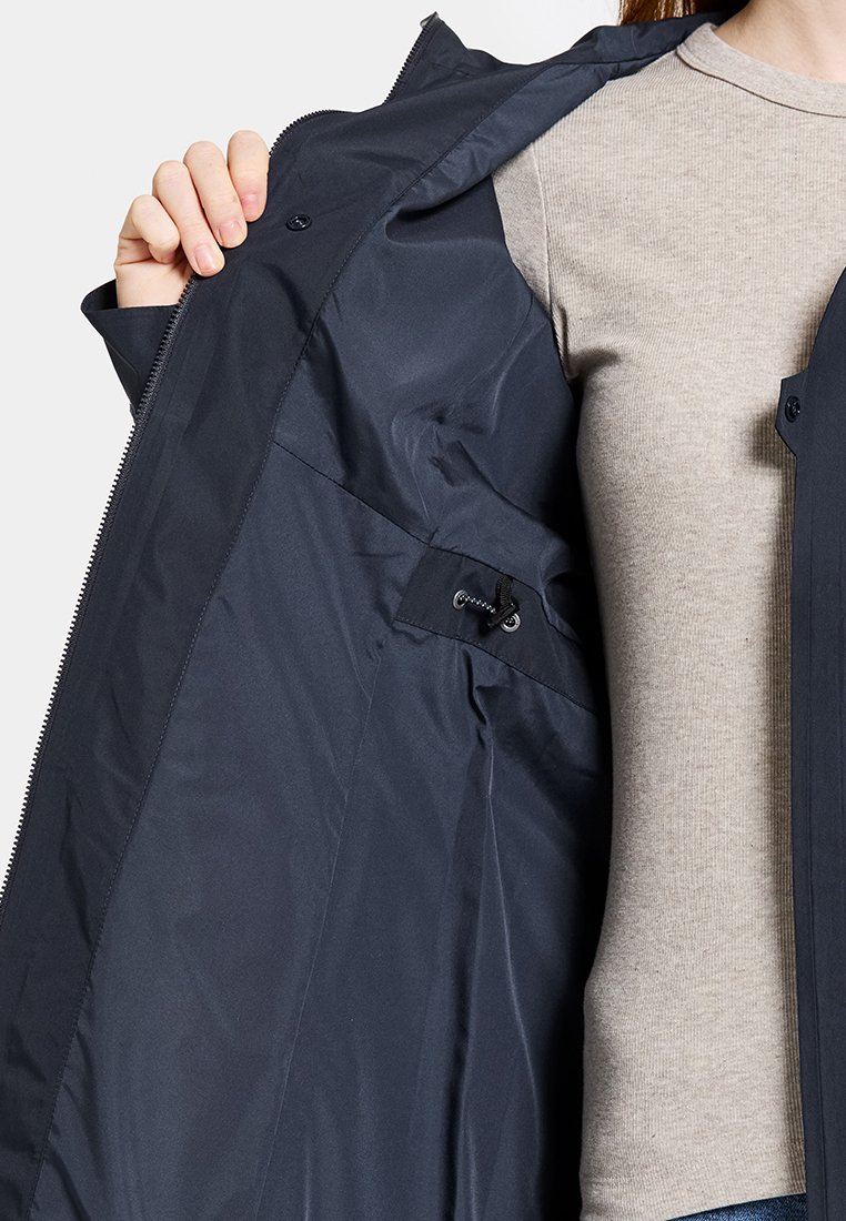 Eine Person hält eine dunkelblaue, wasserdichte Jacke offen, die einen verstellbaren inneren Kordelzug und das glatte Futter der Jacke zeigt.