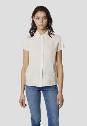 Camisa crema de manga corta con botones delanteros, tela texturizada y bordado floral en el dobladillo. Combinada con jeans de mezclilla azul.